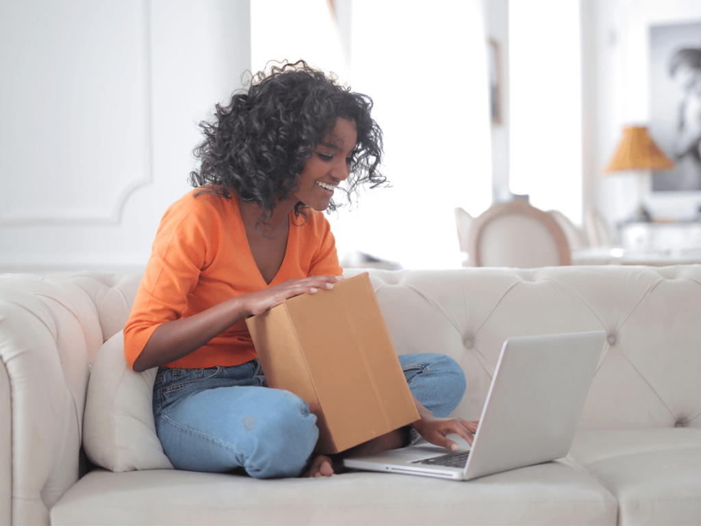 A woman sits on her couch holding a small box, looking at her laptop